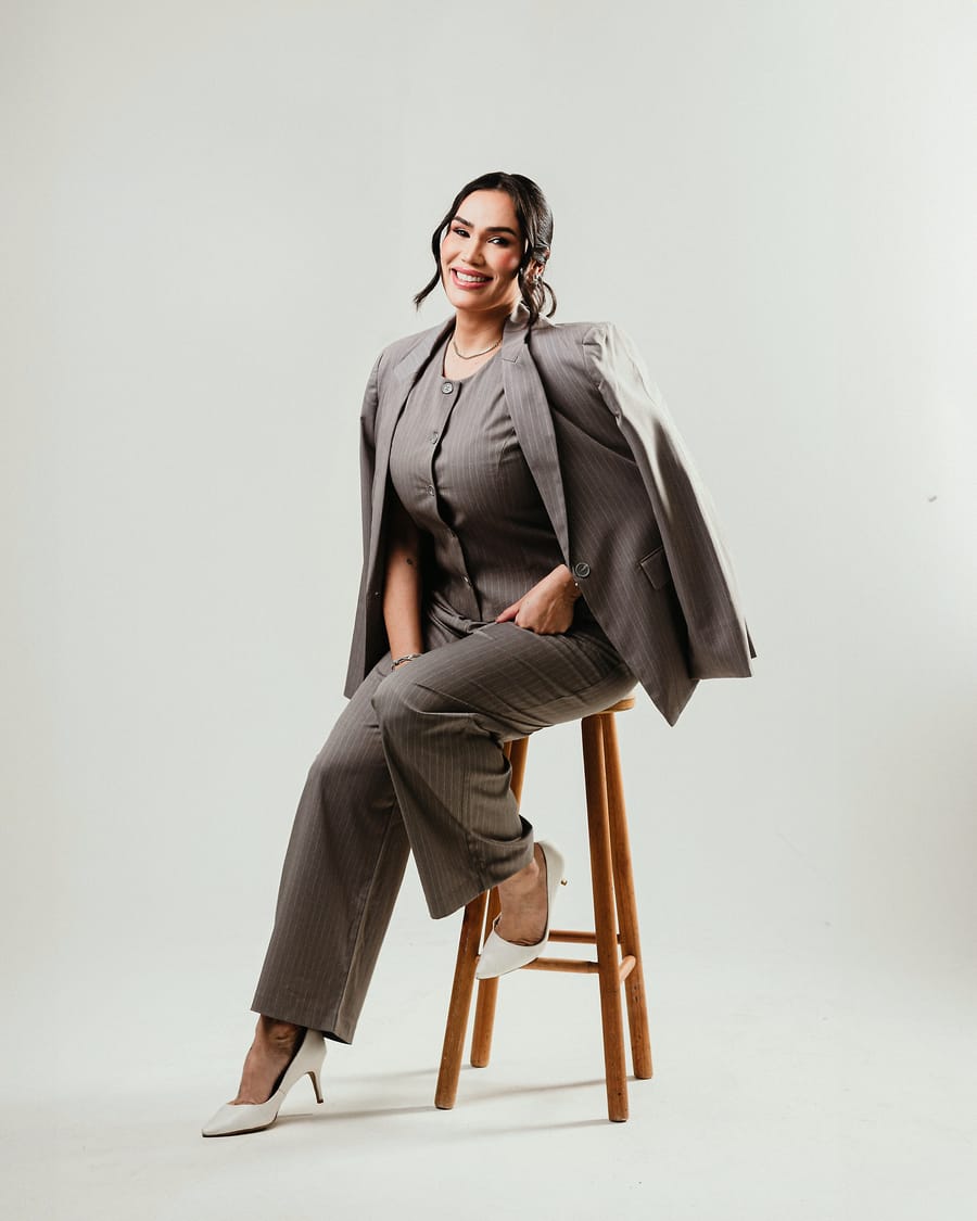 Camila Machado sentada em um banco de madeira, de terno risca de giz, sorrindo